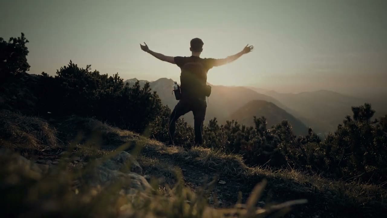 A person on a mountain ridge at sunset, arms open wide.